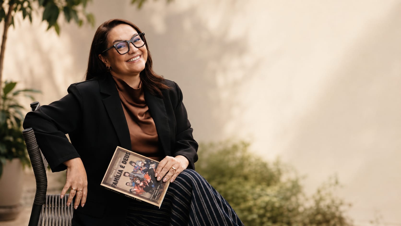 Dra. Karine Schwaab sentada em uma cadeira ao ar livre, de blazer preto, sorrindo e segurando o livro Família é Deus.